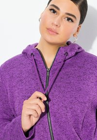 Lila huvtröja med texturerad finish, med en framdragkedja, snoddar och huva. Tyget verkar mjukt och varmt.