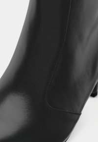 Gros plan sur une botte cheville en cuir noir à texture lisse avec une couture latérale visible, montrant une partie du talon.