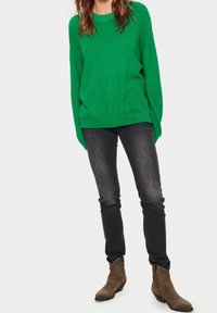 Vihreä neulepaita, jossa on pyöreä kaula-aukko ja alaslasketut olkapäät, yhdistettynä mustiin slim fit -farkkuihin ja ruskeisiin nilkkureihin.