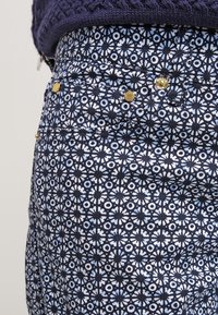 Tejido estampado en azul marino y azul claro con motivos florales. Cuenta con botones de hardware dorado y un acabado texturizado en la cintura.