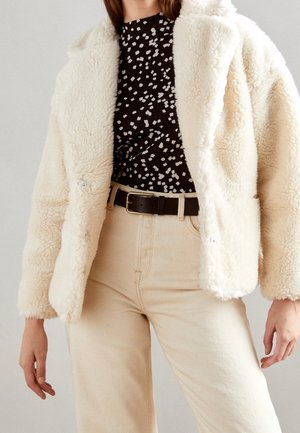Woman wearing cream-colored faux fur jacket, black and white patterned shirt, beige pants, and black belt with gold buckle.