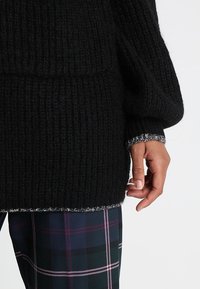 Czarny dziergany sweter z teksturowanym wzorem, z kontrastowymi szarymi wykończeniami na mankietach i dole, zestawiony z kraciastymi spodniami.