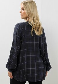 Camicia a quadri blu navy con maniche lunghe e arricciate, caratterizzata da un leggero motivo a scacchi e un'unica spalla posteriore.