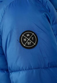 Mėlynas pūkuotas džemperis su lygiu paviršiumi, turintis juodą apvalų pleistrą su geometriniu raštu ir tekstu "MNT 97" centre.