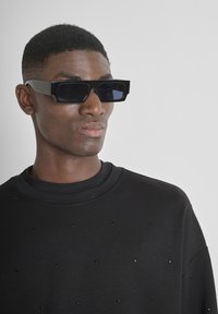 Occhiali da sole rettangolari neri con lenti scure, abbinati a una felpa nera con piccoli dettagli a borchie. Design semplice e moderno.