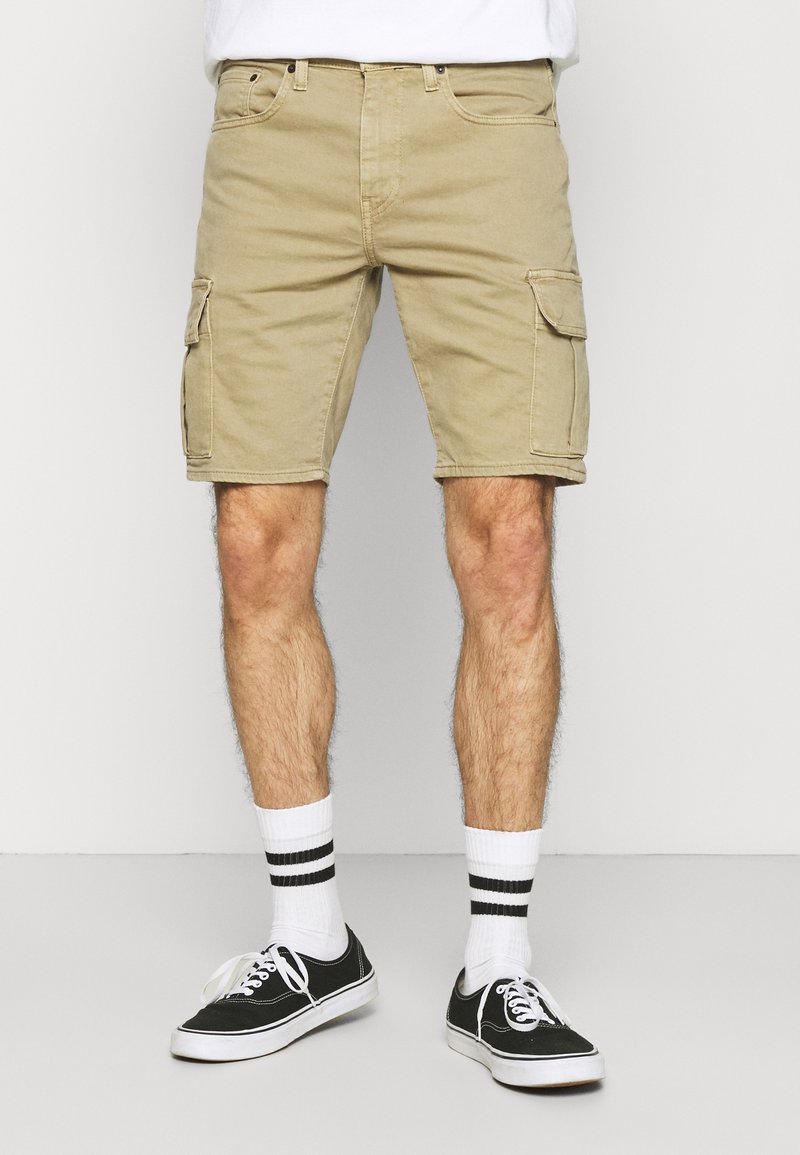 Homme portant un short cargo beige, des chaussettes blanches avec deux bandes noires, et des baskets basses noires avec des lacets blancs.