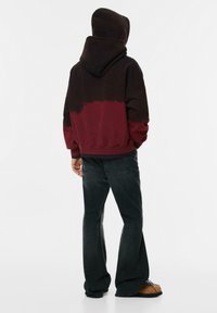 Osoba oblečená v čiernom a burgundskom farebnom bloku mikiny s kapucňou, vo voľných čiernych džínoch a hnedých topánkach, stojí s rukami v vreckách otočená chrbtom.