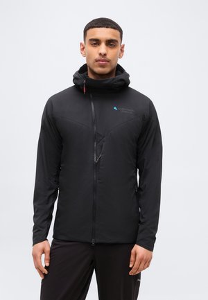 Άνδρας με μαύρο μπουφάν με φερμουάρ, ψηλό γιακά και κουκούλα, στέκεται μπροστά σε απλό ανοιχτό φόντο.