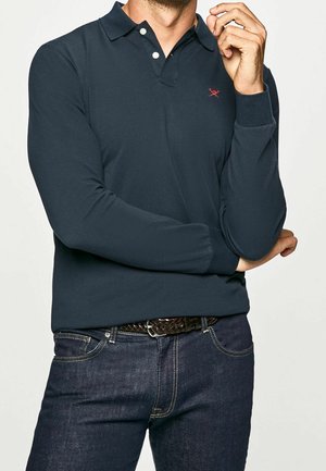 Homme portant un polo bleu foncé à manches longues avec deux boutons, un jean foncé et une ceinture tressée marron, posant avec une main près du visage.
