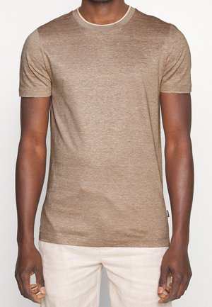 Mann in einem enganliegenden, kurzärmeligen, braun melierten T-Shirt und hellbeigen Hosen, steht vor einem schlichten Hintergrund.