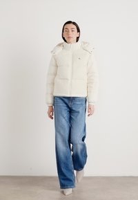 Hvit boblejakke med hette, med glidelås og flate sømmer. Kombinert med lyseblå vide jeans, på en nøytral bakgrunn.