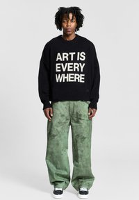 Maglione nero con grande testo bianco "L'ARTE È OVUNQUE", abbinato a pantaloni larghi verdi con texture e scarpe da ginnastica nere.