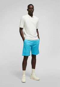 Witte poloshirt met korte mouwen, gecombineerd met blauwe shorts. Model draagt lichtgekleurde sneakers en witte ribgebreide sokken. Eenvoudig ontwerp.