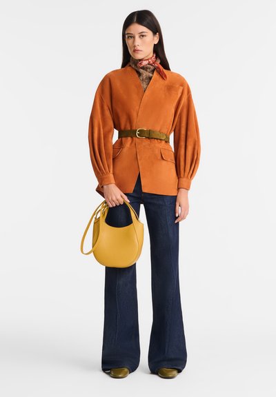 Oranger Wildlederblazer, mit einem grünen Gürtel, kombiniert mit dunkelblauen Schlagjeans. Gelbe strukturierte Handtasche, gemusterter Schal um den Hals.