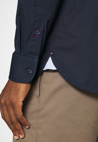 Camicia blu navy con una trama liscia, caratterizzata da un orlo curvo, polsini interni a righe in contrasto e un dettaglio con doppio bottone sui polsini.