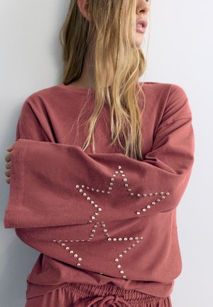 Donna con lunghi capelli biondi indossa una maglietta larga color rosa polvere con disegni di stelle borchiate sulle maniche, in piedi davanti a una parete chiara e semplice.