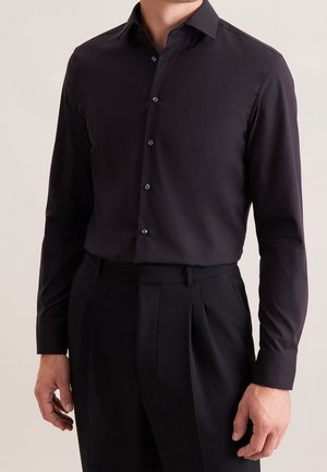 Homme portant une chemise noire ajustée à manches longues boutonnée, rentrée dans un pantalon noir plissé taille haute, sur un fond uni.