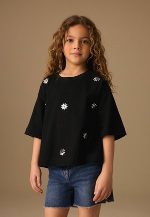 Fiatal lány göndör hajjal, fekete pólót visel ezüst virág díszítéssel és farmer rövidnadrágot, sima háttér előtt áll.