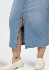 Falda de mezclilla azul claro con una abertura frontal, con un dobladillo sin terminar. Combinada con mocasines blancos sobre un tatuaje de un sol en el tobillo.