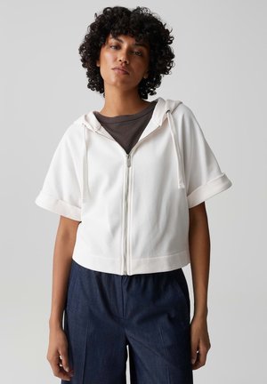 Donna con capelli ricci che indossa una felpa bianca con zip e maniche corte sopra una maglietta scura e pantaloni blu scuro, in piedi davanti a uno sfondo semplice.