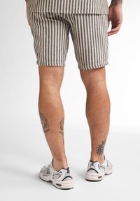 Gestreepte shorts van beige en grijs stof, met een relaxte pasvorm en opgerolde zoom, gecombineerd met witte sneakers. Zichtbare been tattoos.