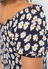 Frau trägt ein dunkelblaues schulterfreies Kleid mit Puffärmeln, das mit weißen und gelben Gänseblümchen-Mustern verziert ist.