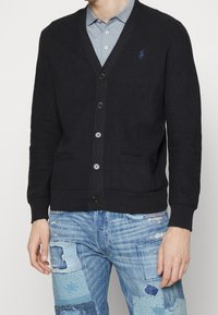 Mörkblå kabelstickad cardigan med V-ringning, sex knappar framtill och två framfickor; har en strukturerad stickning och en liten blå logotyp på bröstet.