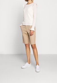 Žena v bílém pleteném topu s dlouhým rukávem a volánky, s béžovými šortkami ke kolenům, v kombinaci s bílými teniskami, stojící na jednobarevném pozadí.