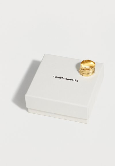 Χρυσό υφασμένο δαχτυλίδι που ακουμπάει σε ένα λευκό κουτί κοσμημάτων Completedworks, πάνω σε απλό λευκό φόντο.