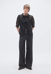 Soprabito in pizzo nero a maniche corte, abbinato a pantaloni in denim scuro a vita alta. Le scarpe sono scure e a punta. Design minimalista, colore monocromatico.