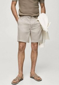 Shorts beige avec une ceinture à cordon ajustable, associés à une chemise décontractée de couleur claire portée sur un bras et des sandales brunes.