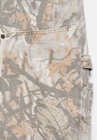 Gros plan d'un tissu avec un imprimé camouflage de feuilles et branches beige, marron clair et gris, avec coutures visibles et détails de poche.