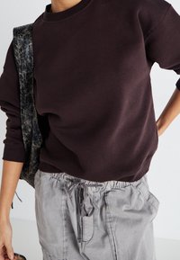 Mörkbrun sweatshirt med rund halsringning och ribbad mudd, bärs tillsammans med ljusgrå cargobyxor med dragsko i midjan. Texturerad tyg synlig.