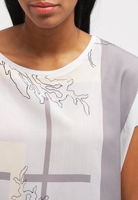 Průhledný top s kulatým výstřihem, zdobený abstraktními liniovými vzory v šedé a béžové barvě na světlém pozadí. Měkká, lehká tkanina.