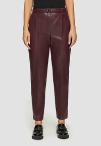 Bordeaux leren broek met een rechte pasvorm, voorzien van een riem in de taille, frontplooien en een gladde textuur. Gdragen met zwarte instapschoenen.