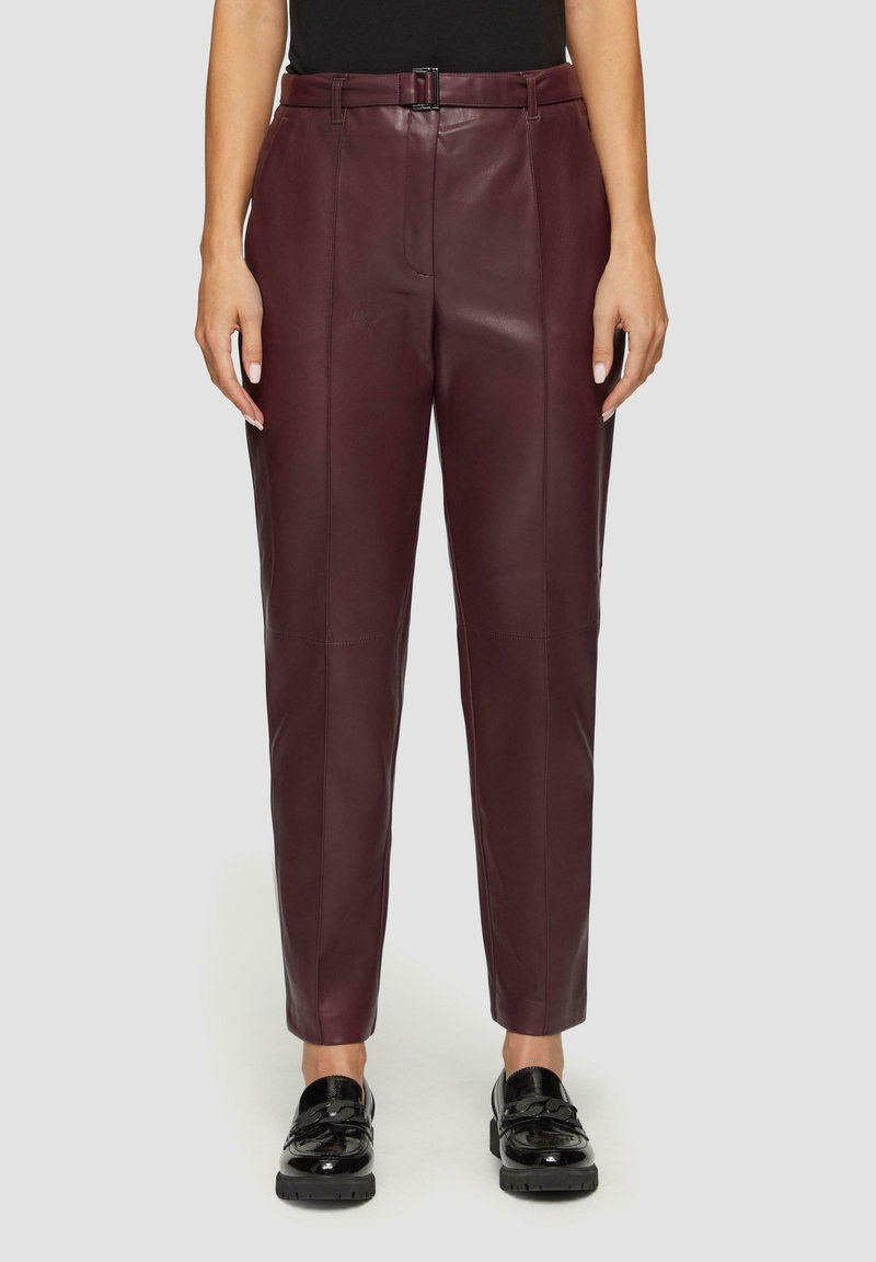 Bordeaux leren broek met een rechte pasvorm, voorzien van een riem in de taille, frontplooien en een gladde textuur. Gdragen met zwarte instapschoenen.