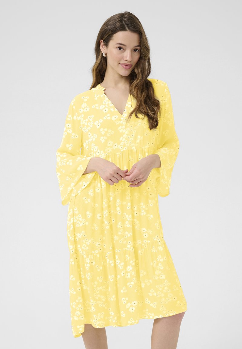 Robe florale jaune avec un décolleté en V, des manches évasées et une jupe à volants. Le tissu a une texture légère avec de petits motifs de fleurs blanches.