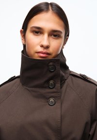 Giovane donna con capelli scuri che indossa un cappotto marrone scuro a collo alto chiuso con tre bottoni, che guarda direttamente davanti a sé su uno sfondo bianco.