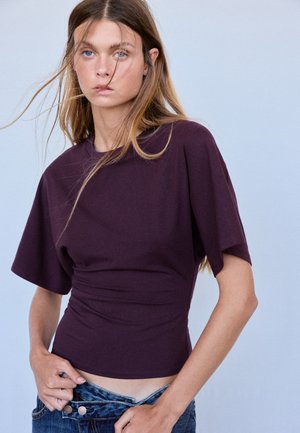 Jeune femme aux longs cheveux porte un haut ajusté violet foncé, ajustant la taille de son jean bleu sur un fond clair uni.