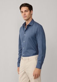 Donkerblauwe blouse met lange mouwen en een button-down kraag, gemaakt van een textuurstof en met knoopsluiting. Gecombineerd met lichtbeige broeken.