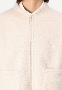 Krämsfärgad fleecejacka med dragkedja framtill, ståkrage och två stora fickor. Den strukturerade tyget ger dimension åt designen.