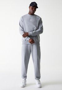 Lys grå sweatshirt og svedbukser lavet af blødt stof, med et broderet logo på brystet og diskret branding på bukserne.