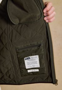 Hand hält eine offene dunkelgrüne gesteppte Jacke und zeigt die innere Reißverschlusstasche mit Joules-Markenetikett und Text auf dem Stoffpatch.