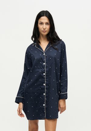 Femme portant une chemise de nuit bleu marine à boutons avec des pois blancs et un liseré blanc sur le col et les poignets, debout devant un fond uni.