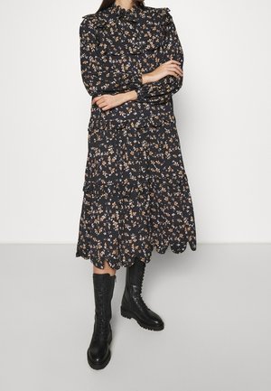 Femme portant une robe midi noire à fleurs avec des volants et un ourlet festonné, associée à des bottes de combat noires à lacets, bras croisés.