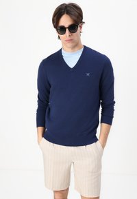 Sotilaslaivaston V-pääntie neule pehmeästä materiaalista, jossa on ribbireunat, päällä vaaleansininen paita ja yhdistettynä kermanvärisiin pinstripe-shorteihin. Mustat aurinkolasit.