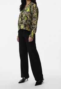 Blouse à manches longues en imprimé léopard vif avec des accents verts et noirs, associée à un pantalon noir ample et des escarpins noirs pointus.