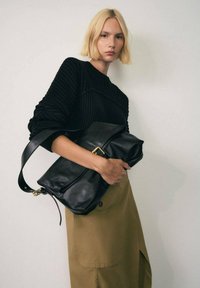 Borsa a mano in pelle nera con una patta piegata, accessori in oro, tenuta sotto il braccio, abbinata a un maglione in maglia nera e una gonna beige. Texture morbida.