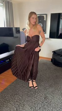 Kvinne i stroppeløs, plissert mørkebrun kjole og svarte hæler som står i stuen og holder i kjolens skjørt.