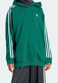 Grön huvtröja med vita sidostråk, en framficka och en broderad logotyp på bröstet. Tillverkad av mjukt, texturerat material.